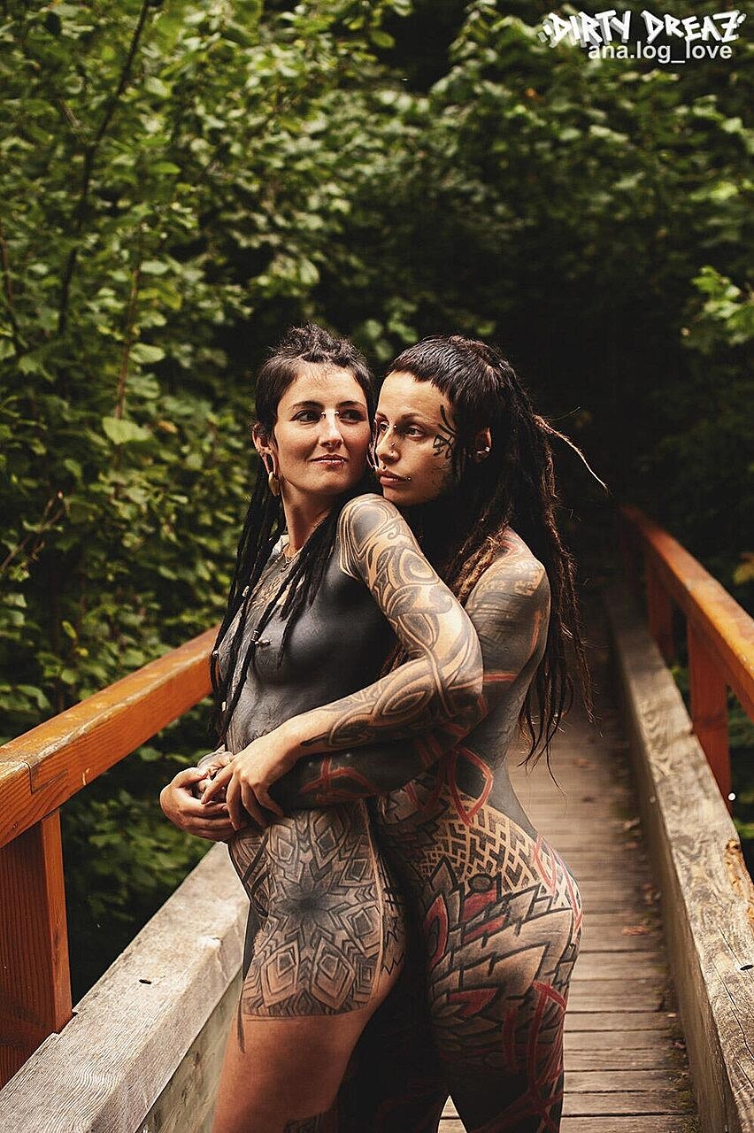 Heavily tattooed lesbians hold each other while totally naked on a bridge